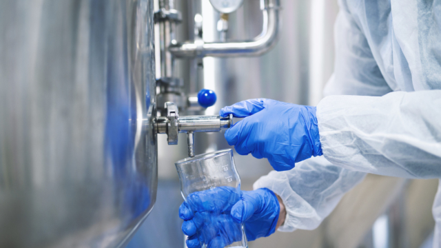 Close up view of hands in protective gloves opening tap valve and filling glass with samples from industrial machine reservoir.